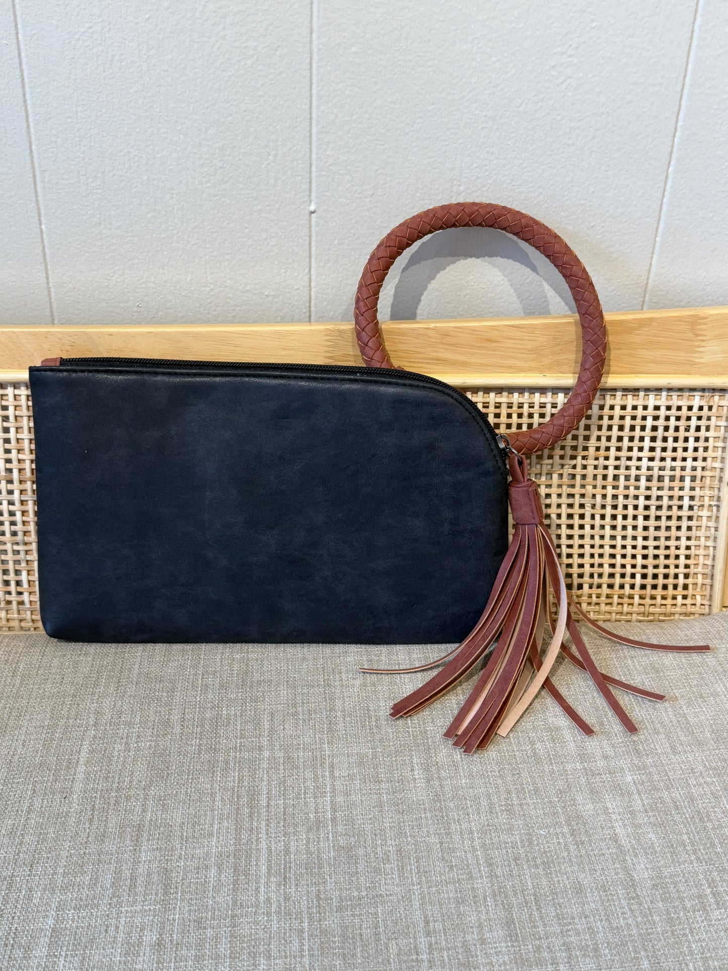 A black suede clutch bag with a circular brown leather handle and matching leather tassels, displayed on a woven cane panel against a light gray wall.