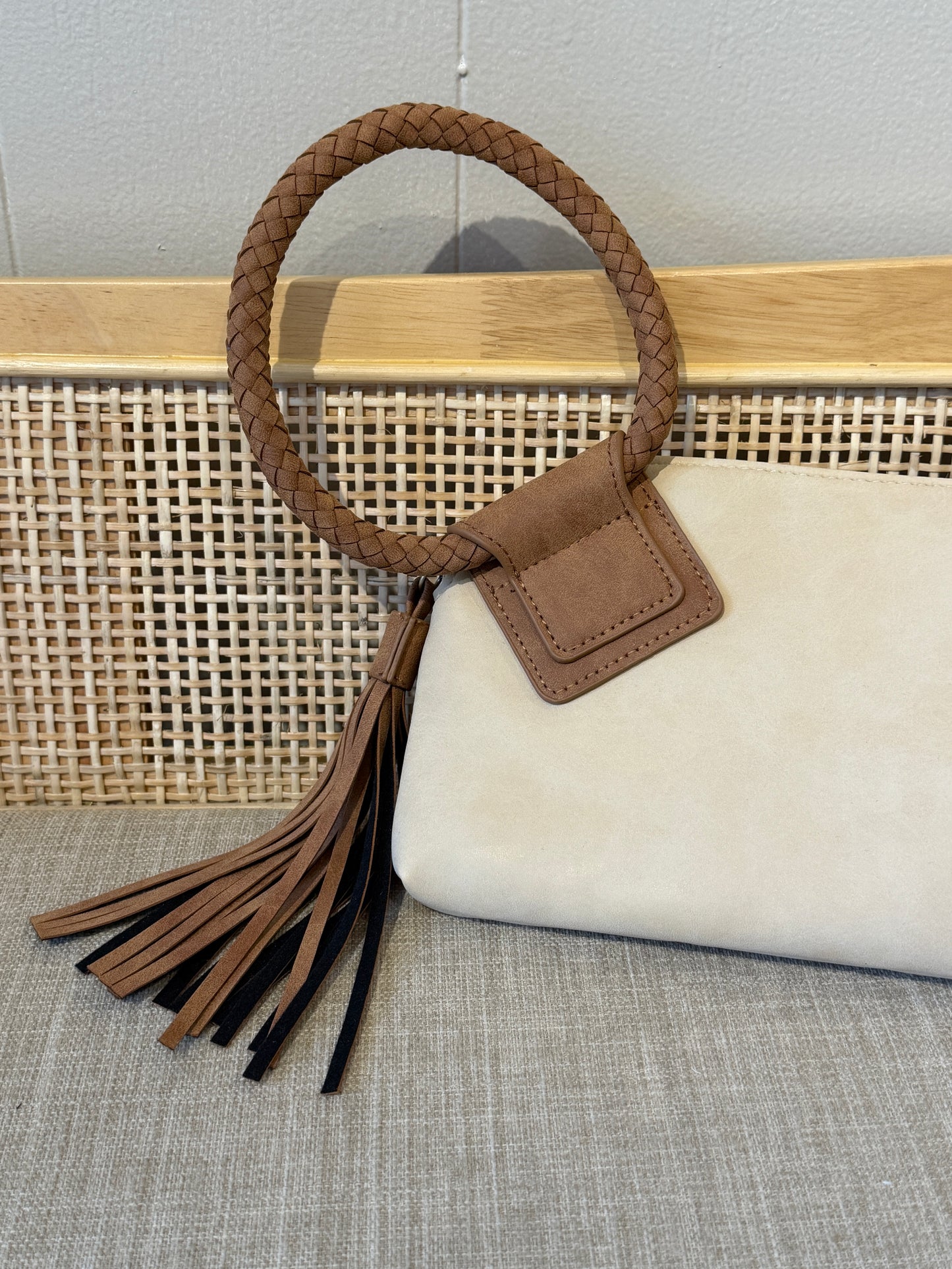 A cream-colored suede clutch bag with a brown braided leather handle and tassel, displayed on a woven cane chair against a light gray wall.