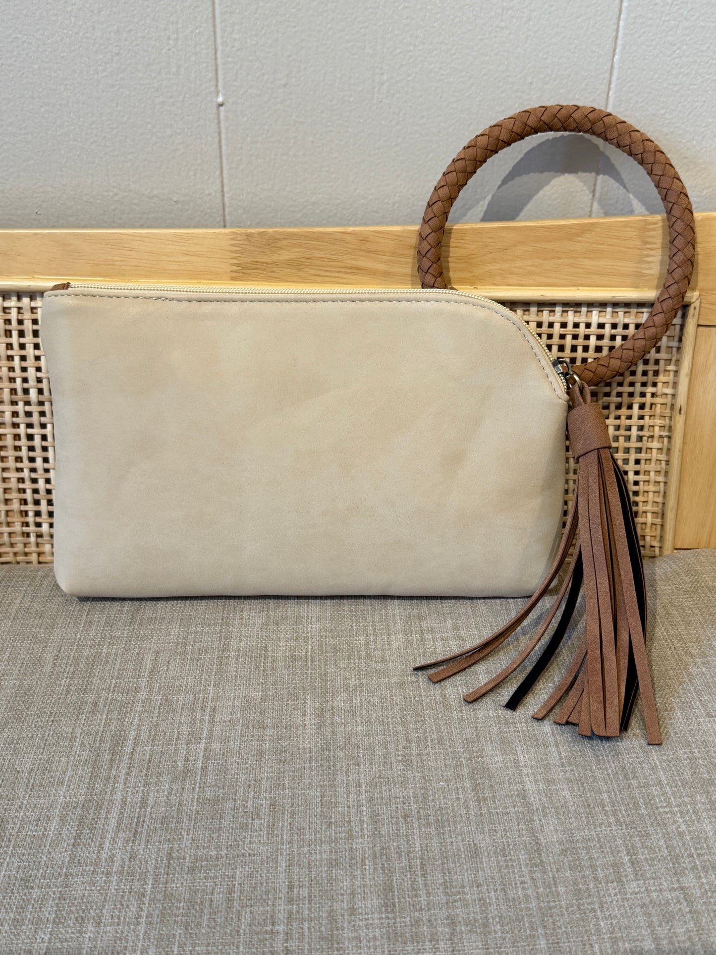 A beige suede clutch bag with a brown braided leather handle and matching tassel, displayed on a woven cane bench against a light gray wall.