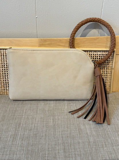 A beige suede clutch bag with a brown braided leather handle and matching tassel, displayed on a woven cane bench against a light gray wall.