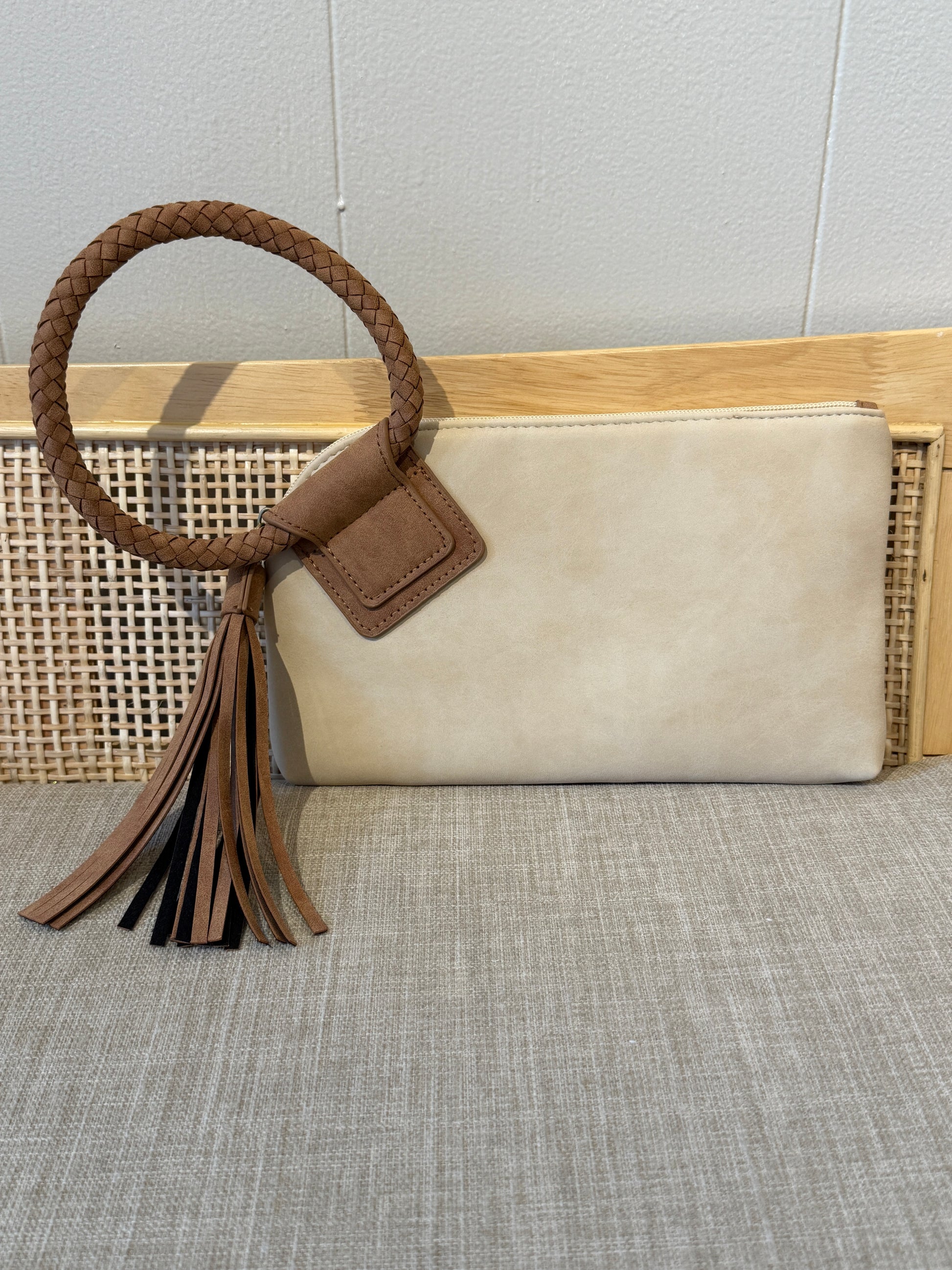 A cream-colored faux suede clutch bag with a brown braided leather handle and a matching brown leather tassel, displayed on a woven cane bench against a light gray wall.