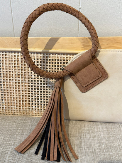 A ivory leather handbag with a large tan braided oval handle, a tassel detail, and a small rectangular leather accent, displayed against a woven cane panel and light wood background.