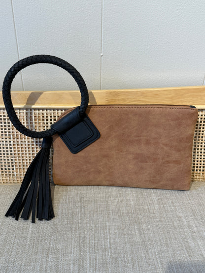 A brown suede clutch bag with a black braided rope handle and a black leather tassel, displayed on a woven cane bench against a light gray wall.