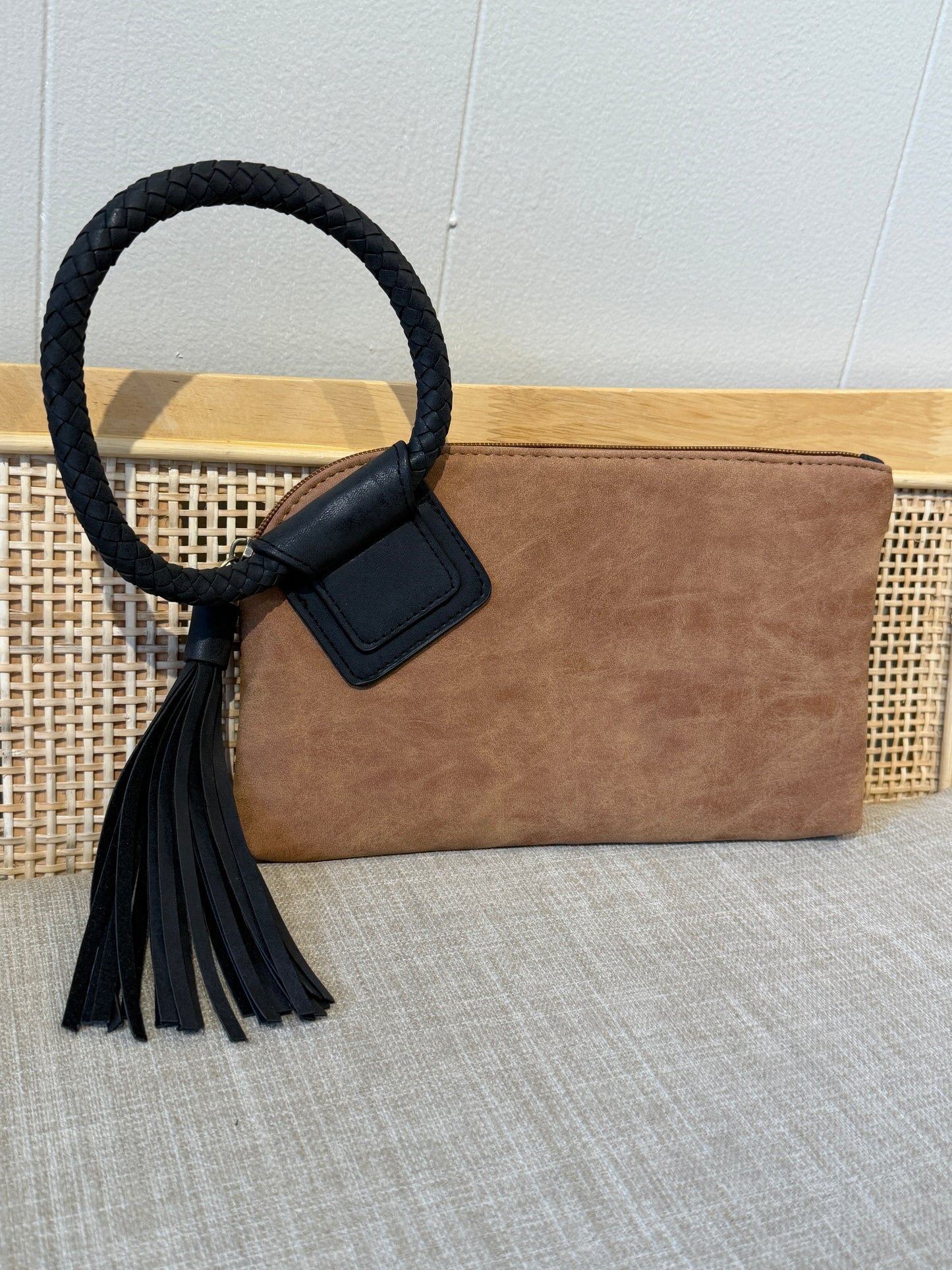 A brown suede clutch bag with a black braided rope handle and a black leather tassel, displayed on a woven cane panel against a light gray wall.