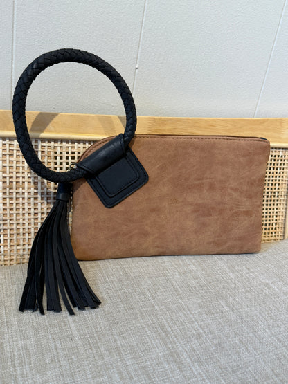 A brown suede clutch bag with a black braided rope handle and a black leather tassel, displayed on a woven cane panel against a light gray wall.