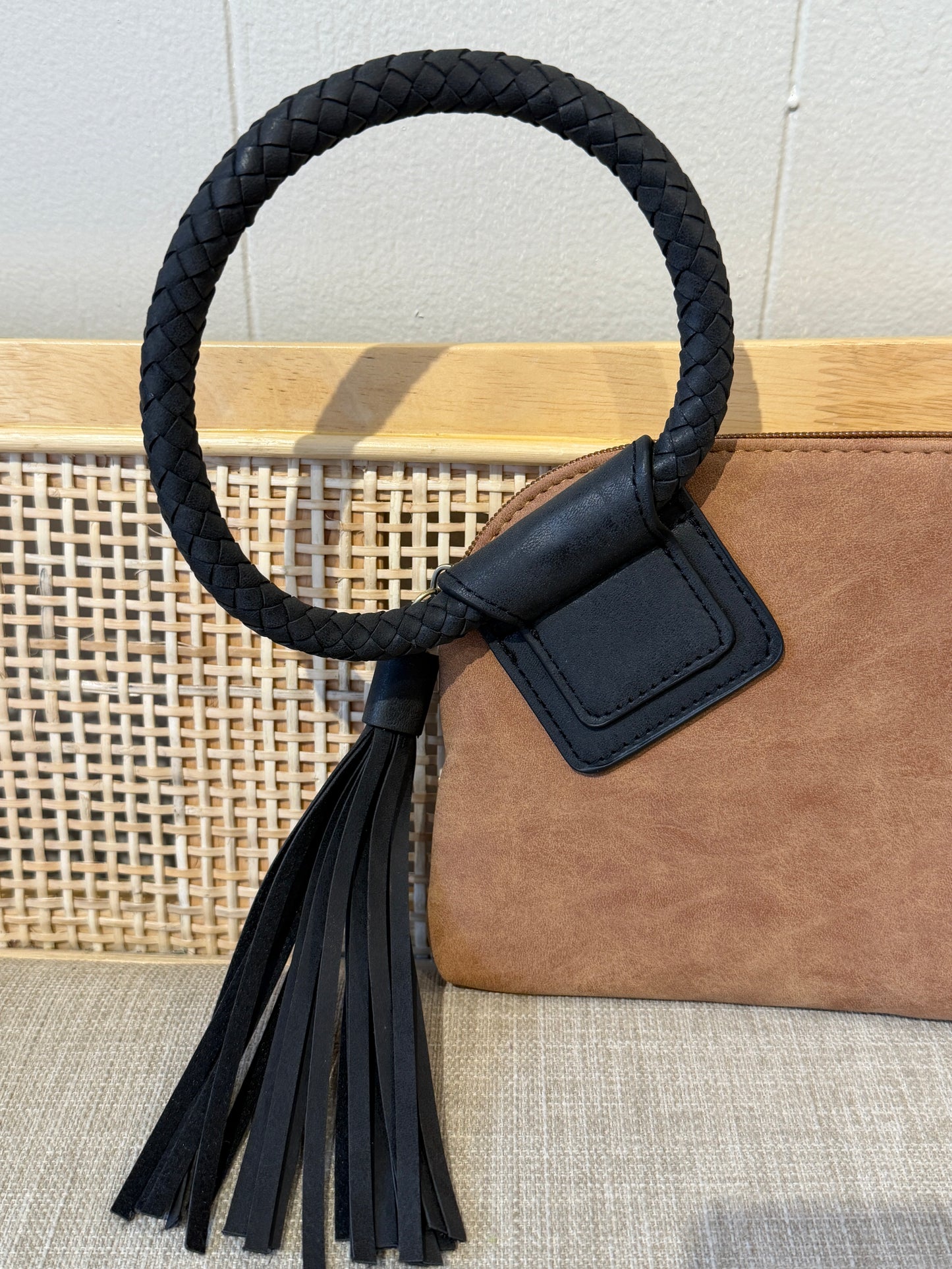 A close-up of a brown leather handbag featuring a large black braided circular handle with a black leather tassel and a small black leather tag, set against a light wood cane panel and beige fabric surface.
