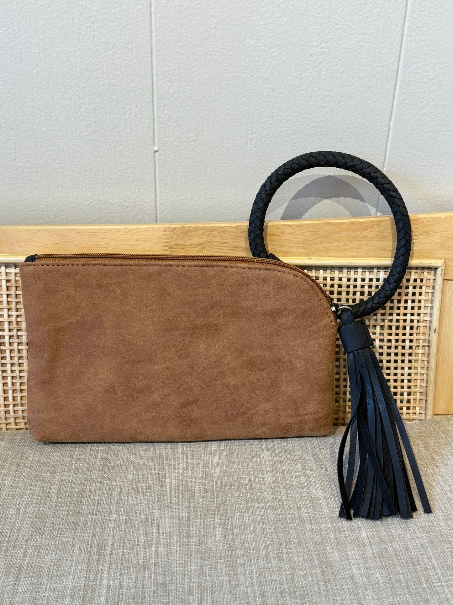 A brown faux-leather clutch bag with a black braided rope handle and a matching black tassel, displayed on a woven cane panel and textured beige fabric surface.