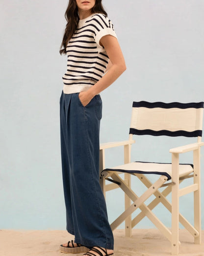 Woman wearing a striped short sleeve cream and black knit sweater and blue pants standing on sand with a chair in the background.