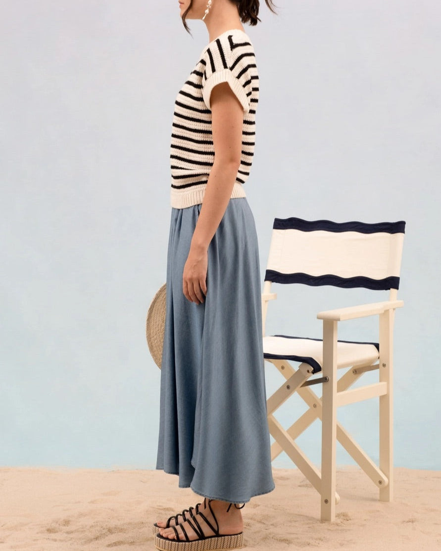 Woman wearing a striped short sleeve cream and black knit sweater and blue skirt standing on sand with a chair in the background.
