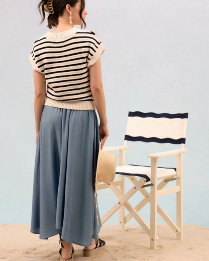 Woman wearing a striped short sleeve cream and black knit sweater and blue skirt standing on sand with a chair in the background.