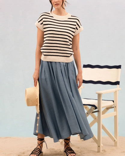 Woman wearing a striped short sleeve cream and black knit sweater and blue skirt standing on sand with a chair in the background. 
