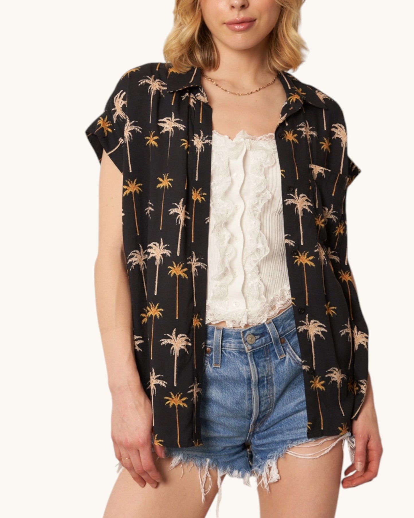 Woman wearing a black palm tree patterned shirt over a white top and denim shorts on a white background.
