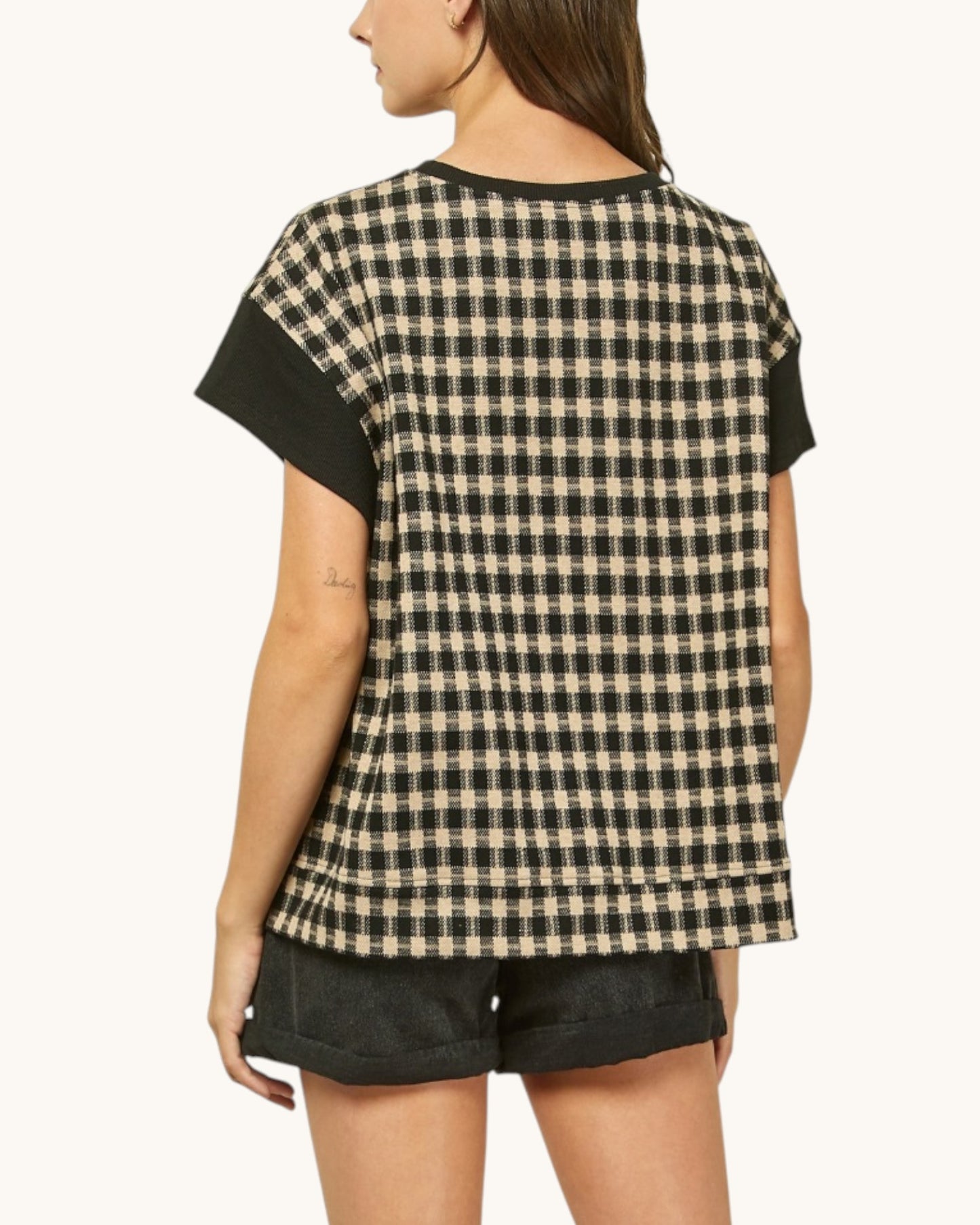 A woman shown from the back wearing a short-sleeve black and beige gingham patterned top with solid black sleeves, paired with black denim shorts, against a plain off-white background.