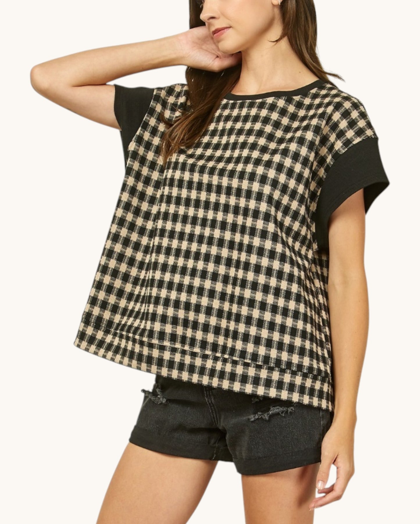 A woman wearing a short-sleeve top with a black and beige gingham pattern and solid black sleeves, paired with black denim shorts, against a plain off-white background.