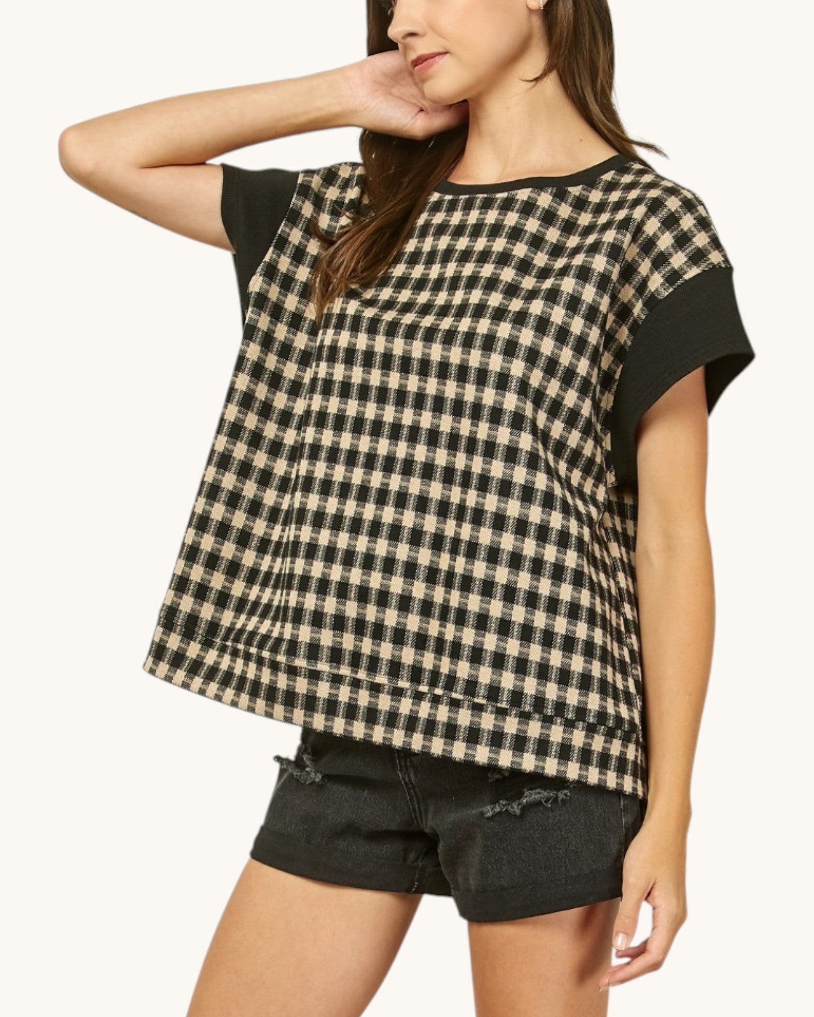A woman wearing a short-sleeve top with a black and beige gingham pattern and solid black sleeves, paired with black denim shorts, against a plain off-white background.