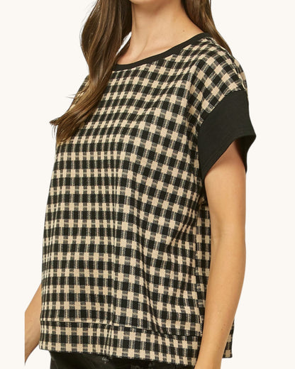 A woman wearing a short-sleeve top with a black and beige gingham pattern and solid black sleeves, shown against a plain light background.