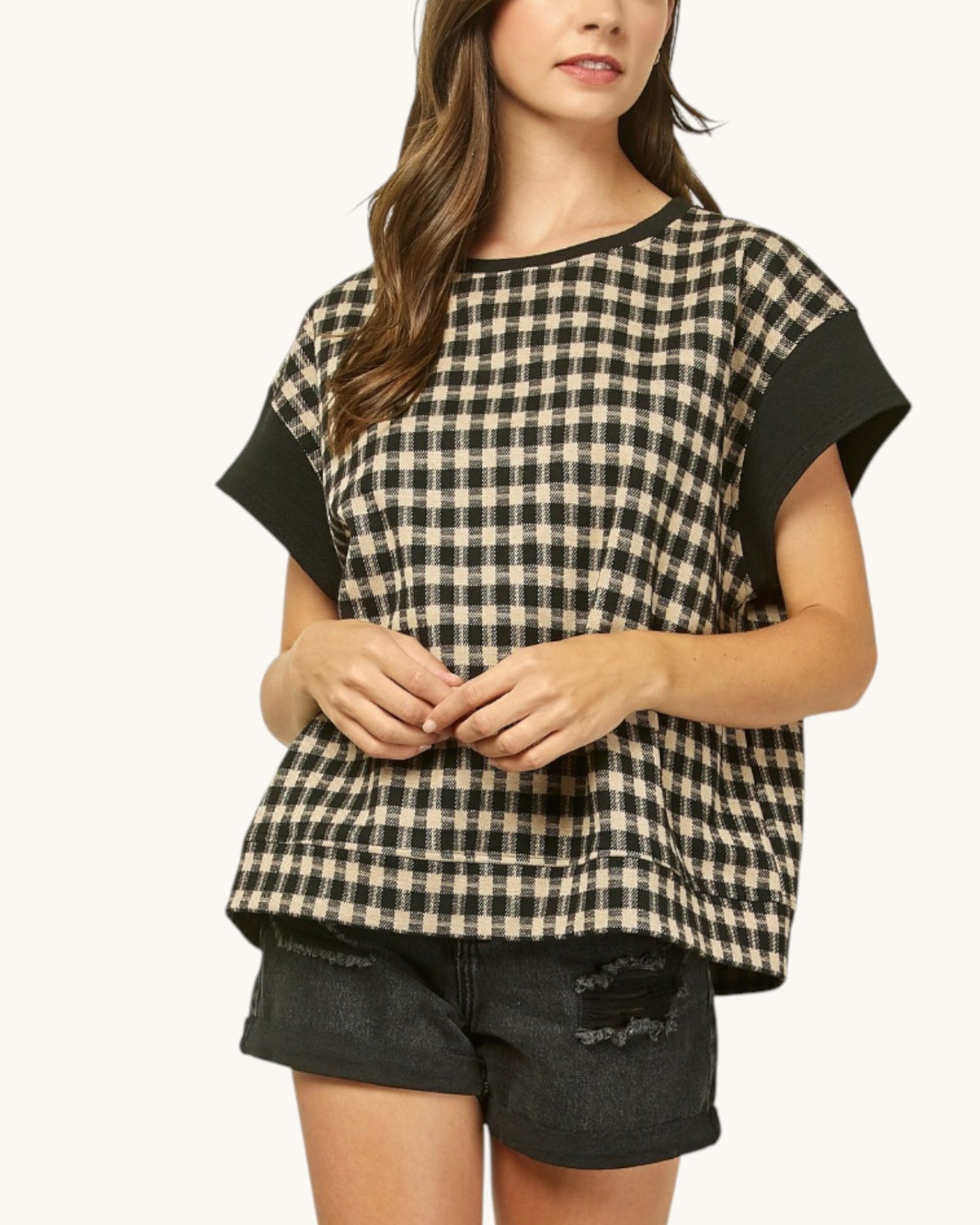 A woman wearing a black and beige gingham patterned short-sleeve top with solid black sleeves, paired with black denim shorts, against a plain off-white background.