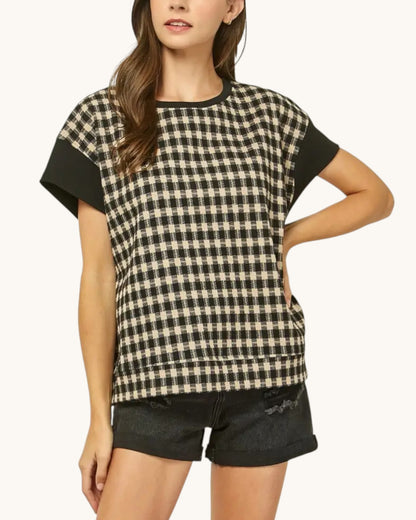 A woman wearing a short-sleeve black and beige gingham patterned top with solid black sleeve panels and black denim shorts, photographed against a plain light background.