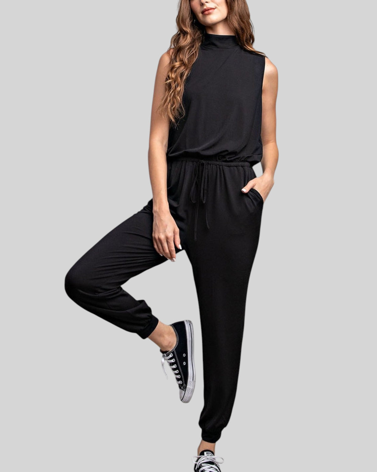 A woman modeling a black sleeveless mock-neck jumpsuit with a drawstring waist and relaxed jogger-style pants, paired with black sneakers, against a solid light gray background.