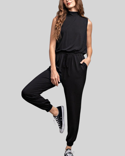 A woman modeling a black sleeveless mock-neck jumpsuit with a drawstring waist and relaxed jogger-style pants, paired with black sneakers, against a solid light gray background.