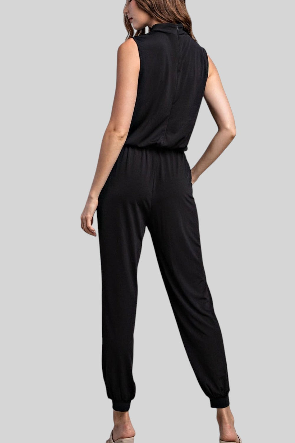 A woman shown from the back wearing a black sleeveless jumpsuit with a high collar and relaxed tapered legs, photographed against a plain light gray studio background.