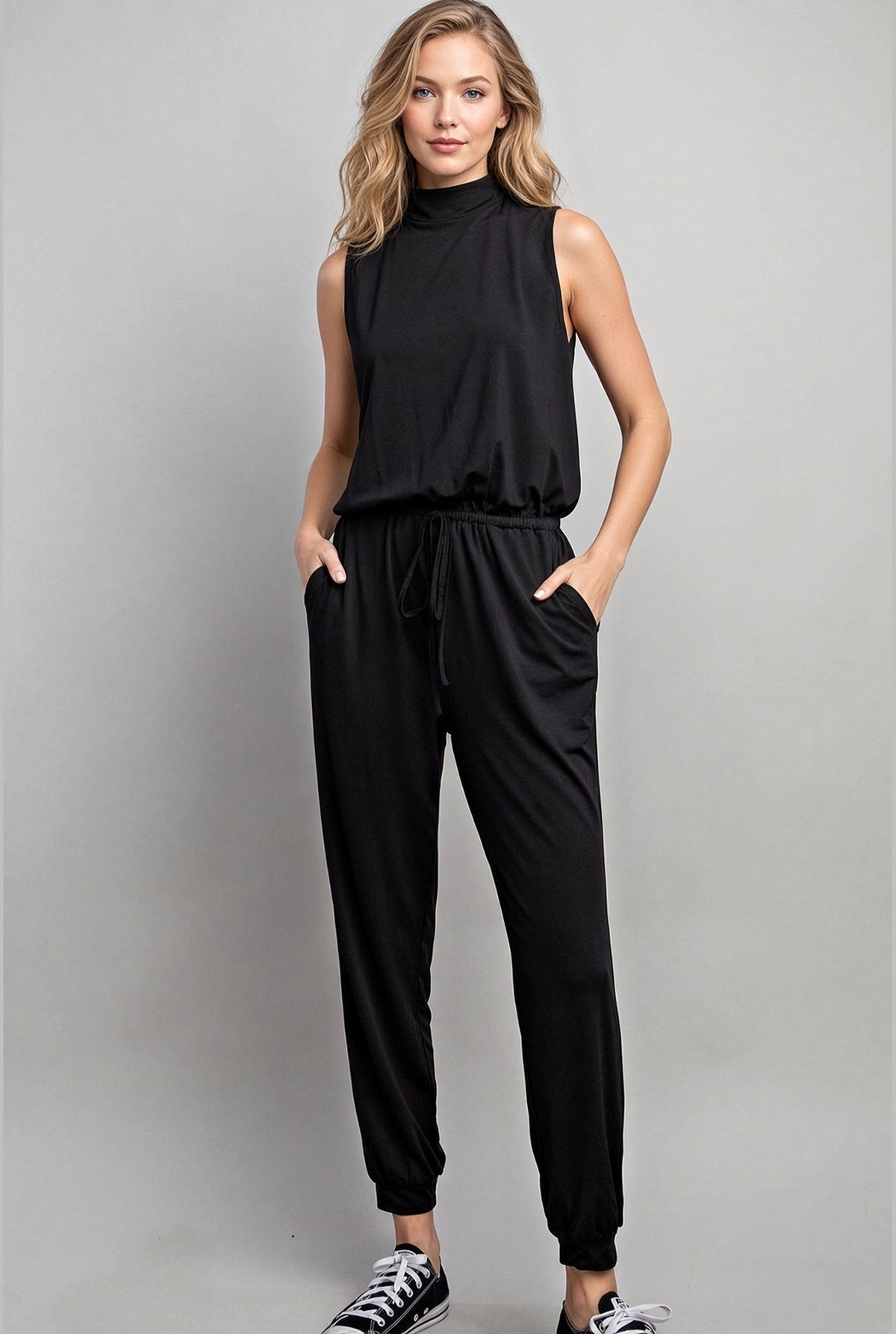 Woman wearing a silky sleeveless black mock neck jumpsuit and black sneakers.