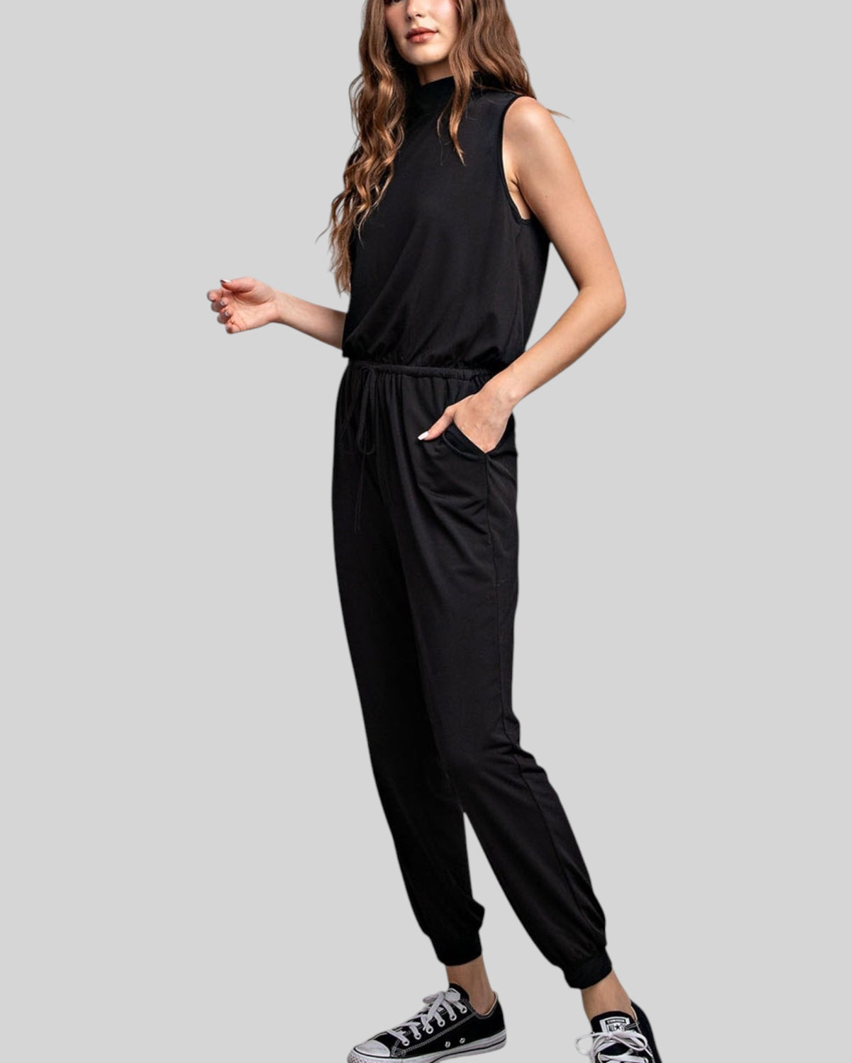 A woman modeling a black sleeveless mock-neck jumpsuit with side pockets and elastic jogger-style legs, paired with black sneakers, against a solid gray background.