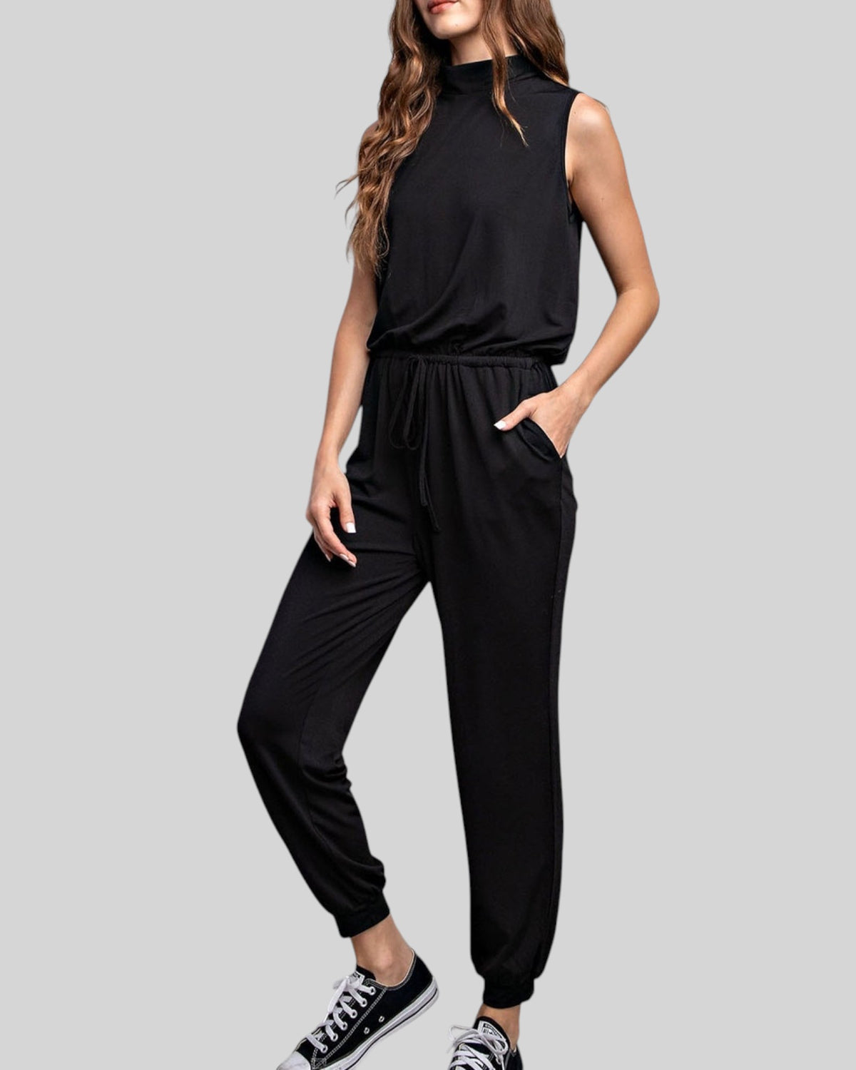 A woman modeling a black sleeveless mock-neck jumpsuit with a drawstring waist and side pockets, paired with black sneakers, against a solid gray background.