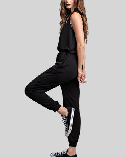 A woman modeling a black sleeveless top and black jogger pants, posed against a solid light gray background.