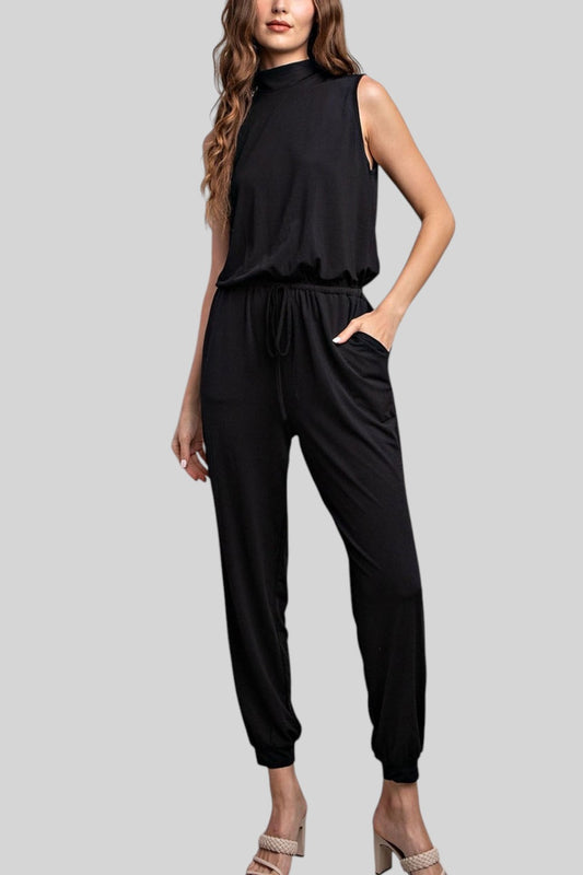 A woman modeling a sleeveless black mock-neck jumpsuit with a drawstring waist and relaxed jogger-style legs, shown against a plain light gray studio background.