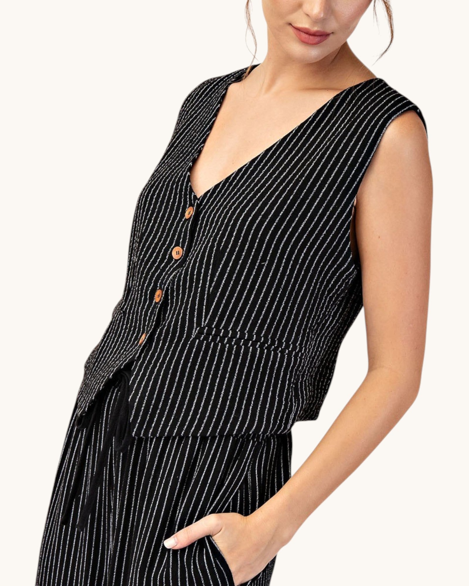 Side view of a woman wearing a cropped, fitted black pinstripe sleeveless button up vest and matching pants. 