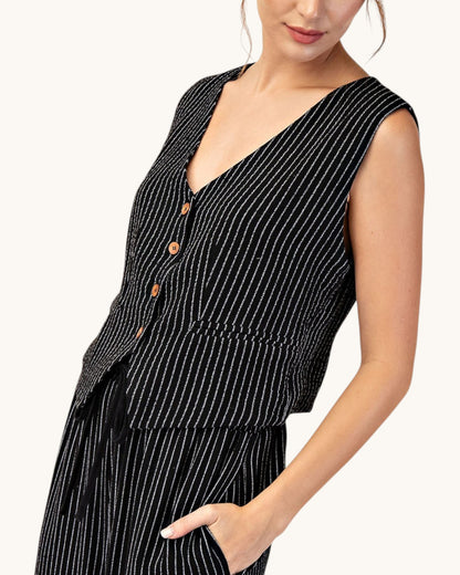 Side view of a woman wearing a cropped, fitted black pinstripe sleeveless button up vest and matching pants. 