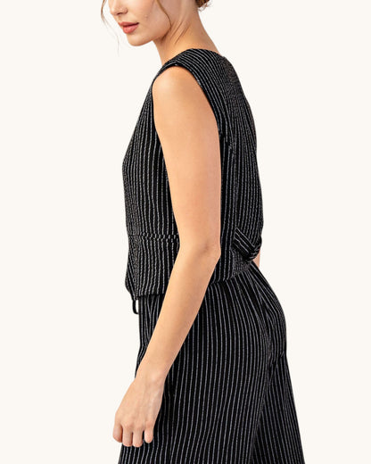 Side and back view of a woman wearing a cropped, fitted black pinstripe sleeveless button up vest and matching pants. 