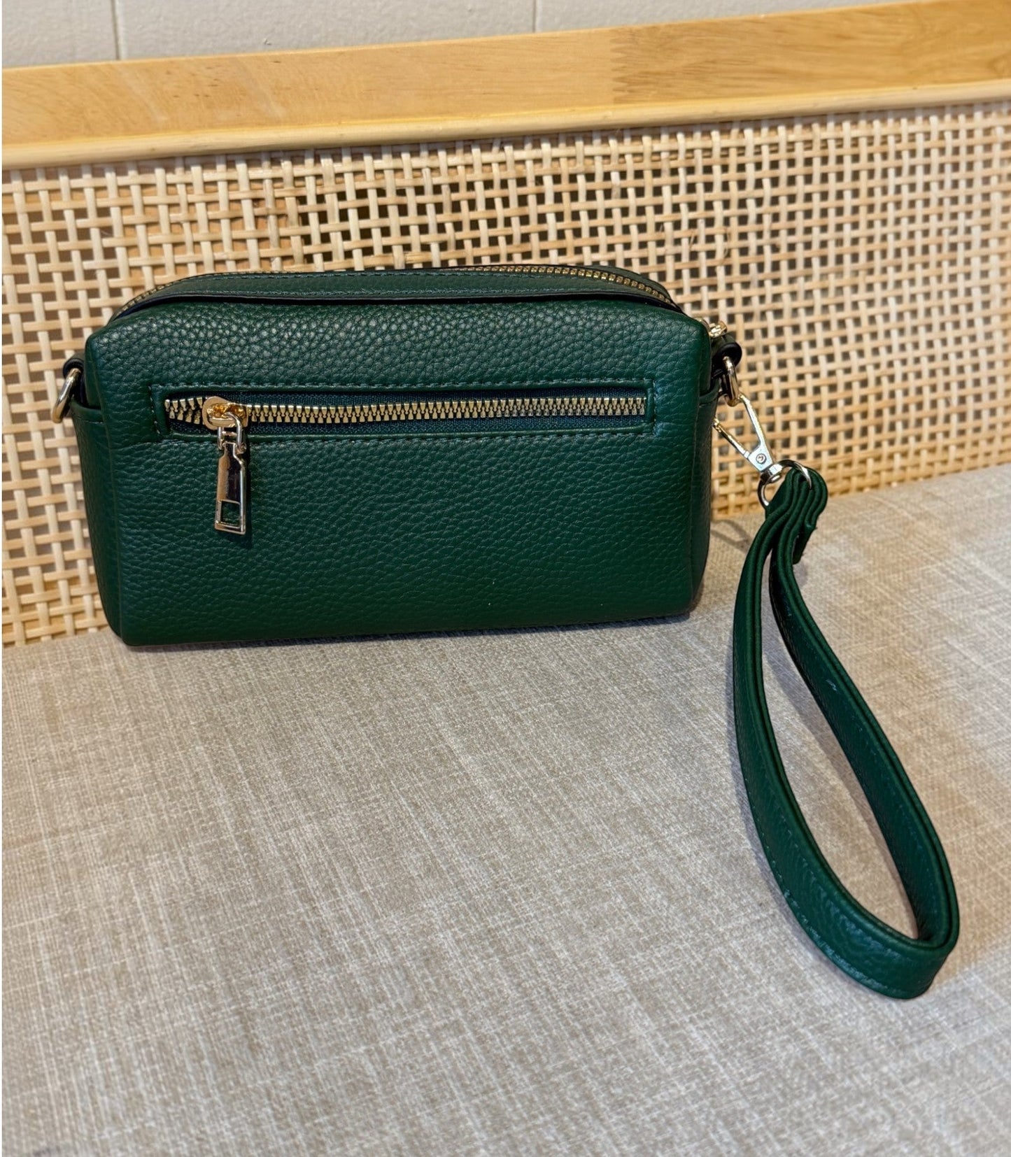 A dark green pebbled leather wristlet clutch bag with a gold zipper and a matching green wrist strap, resting on a textured beige fabric surface in front of a woven cane panel.