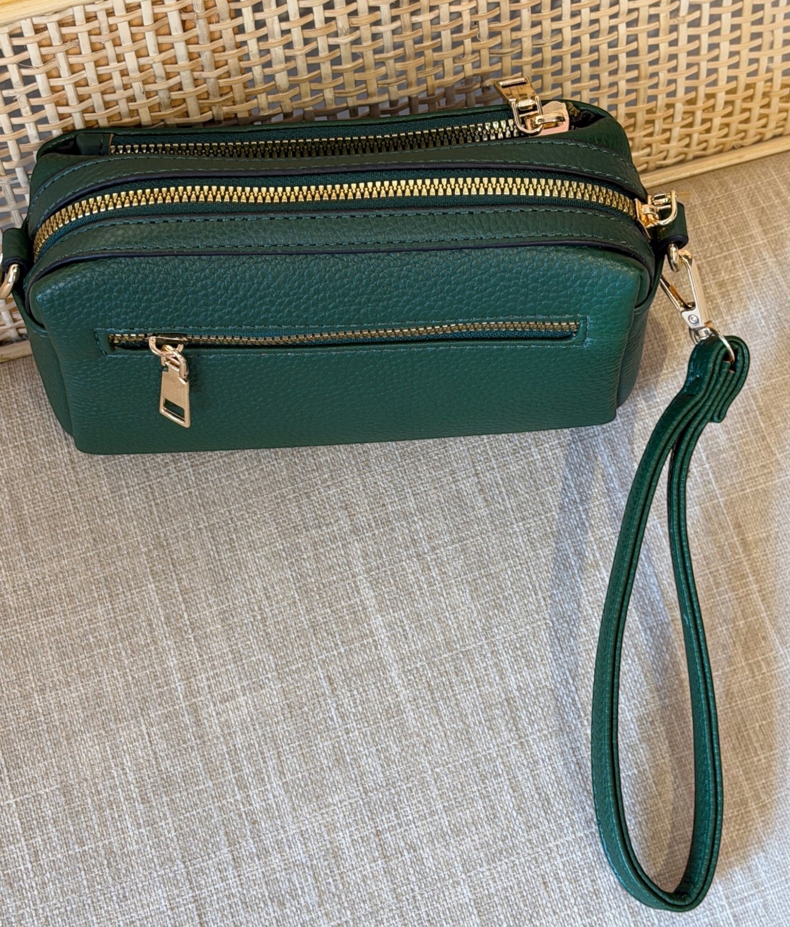 A dark green pebbled leather crossbody bag with gold zippers and a matching wristlet strap, displayed on a textured beige fabric surface.