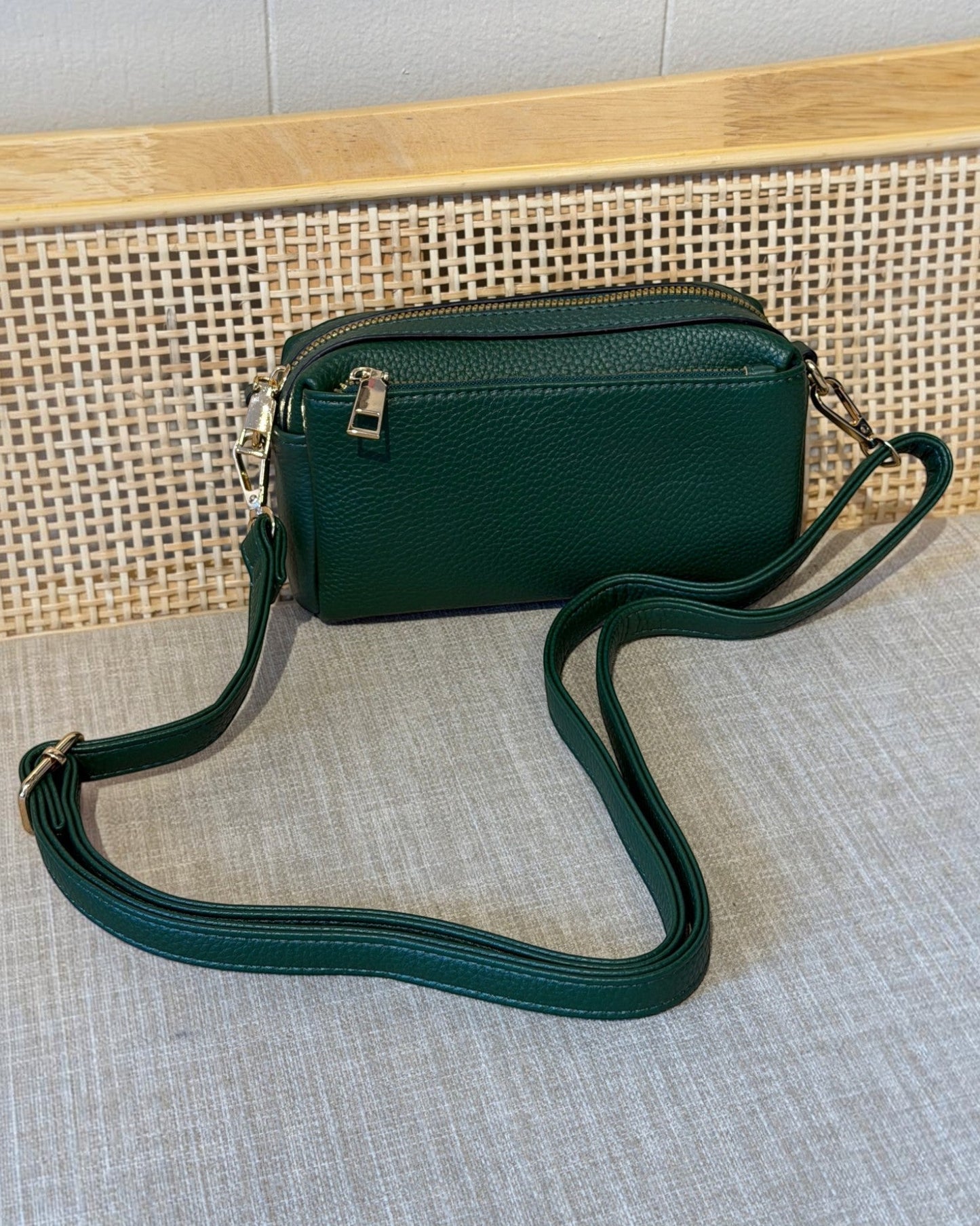 A dark green pebbled leather crossbody bag with a matching adjustable shoulder strap and gold-tone hardware, displayed on a textured gray fabric surface in front of a woven cane panel and light wood background.
