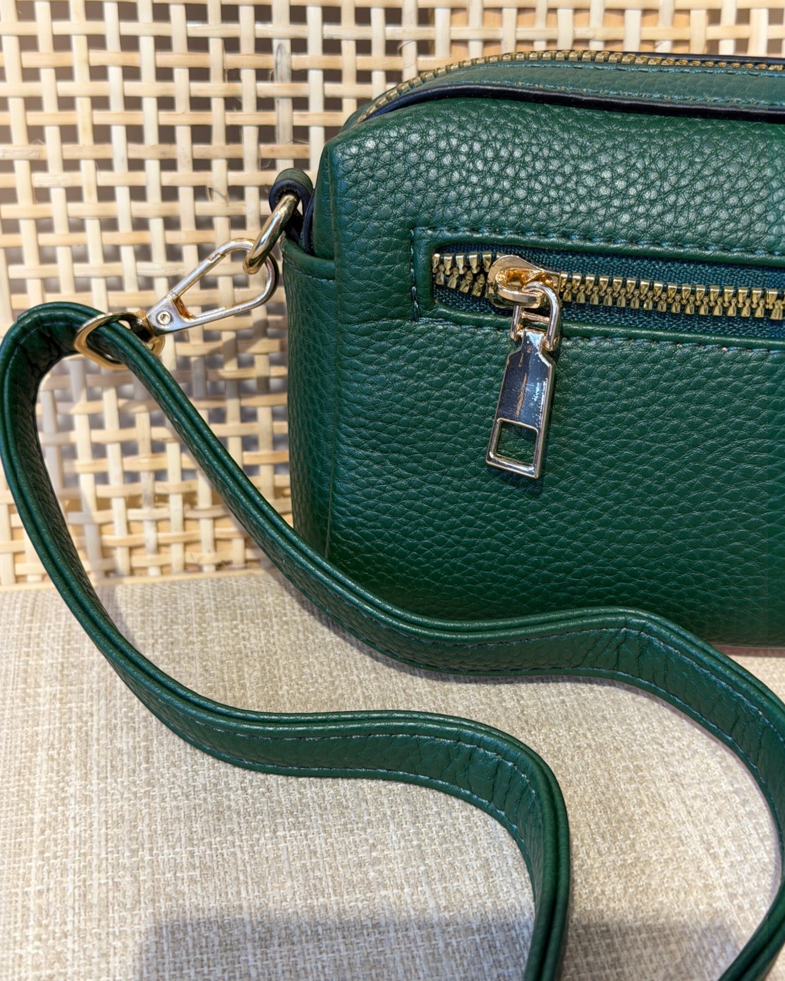 A close-up of a dark green pebbled leather crossbody bag with a matching strap, gold zipper, and gold-tone hardware, resting on a textured beige surface.