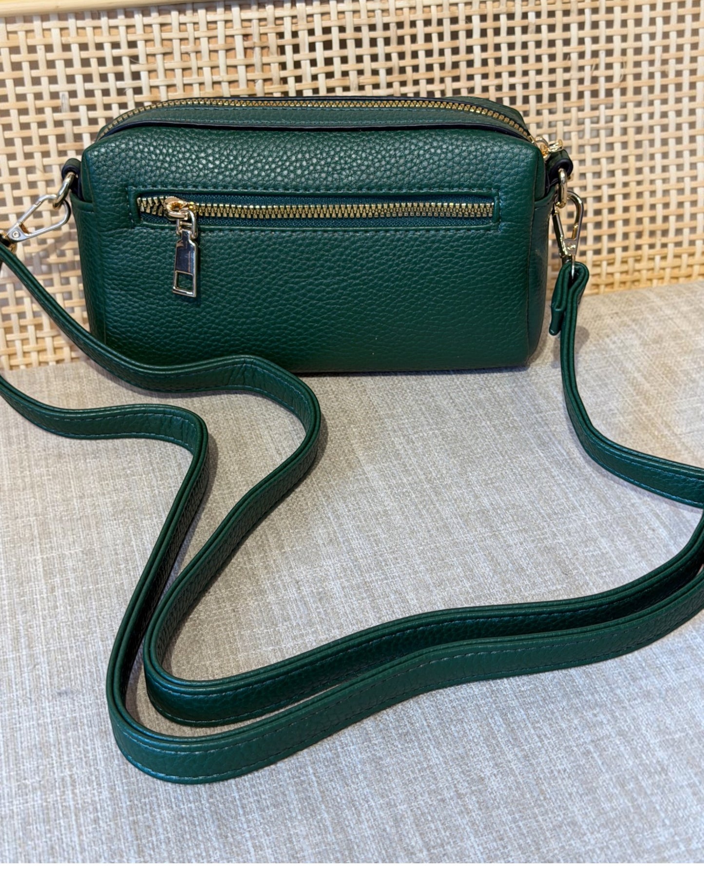 A dark green pebbled leather crossbody bag with a gold zipper and a matching green strap, displayed on a textured beige fabric surface.