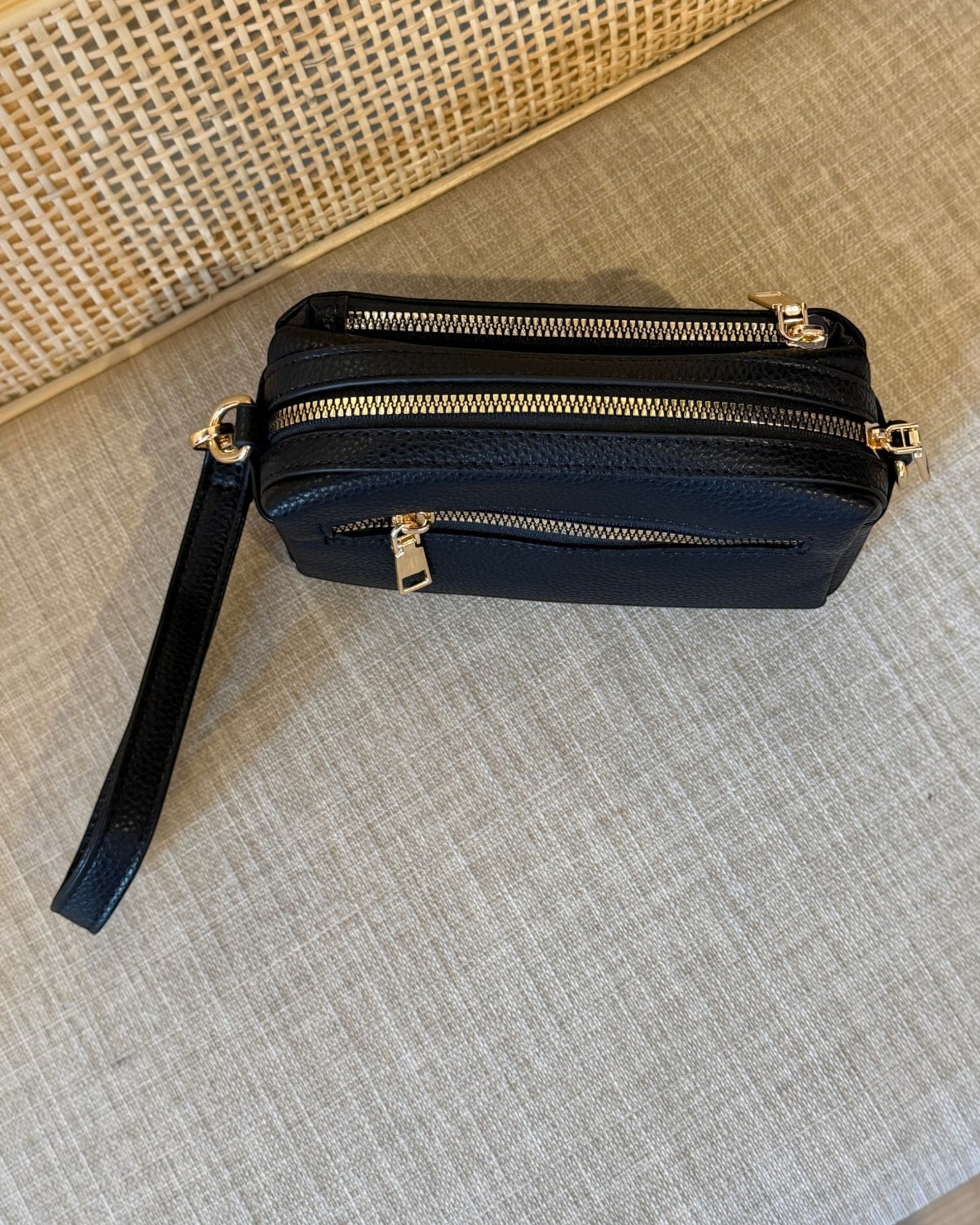 A black pebbled leather crossbody bag with gold-tone hardware, featuring three horizontal zippered compartments and a wristlet strap, resting on a textured beige fabric surface.