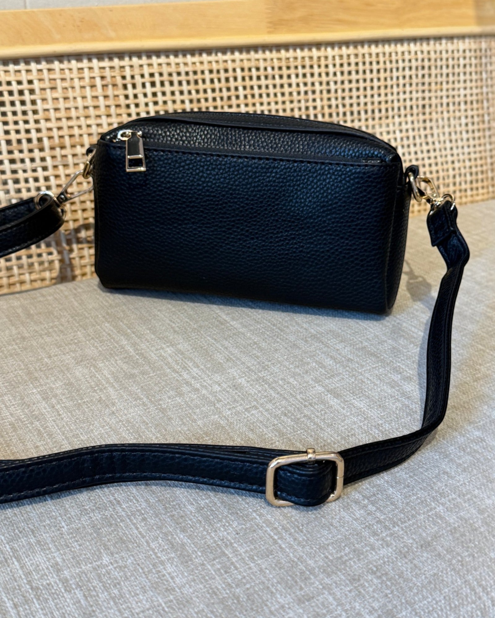 A black pebbled leather crossbody bag with a top zip closure and a matching adjustable shoulder strap, resting on a textured light gray fabric surface in front of a woven cane screen.