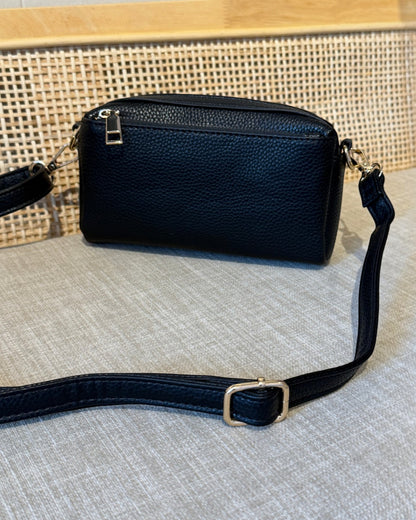 A black pebbled leather crossbody bag with a top zip closure and a matching adjustable shoulder strap, resting on a textured light gray fabric surface in front of a woven cane screen.