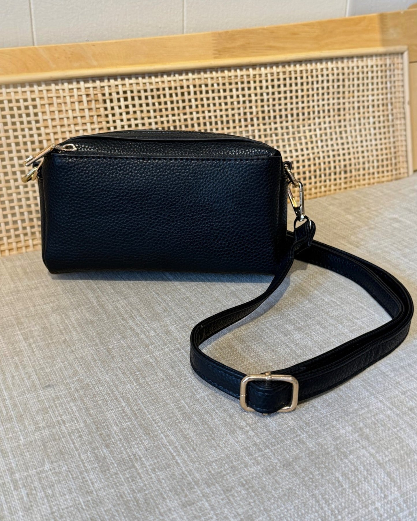 A black pebbled leather crossbody bag with a matching adjustable strap and gold-tone hardware, displayed on a light gray textured surface with a woven cane panel in the background.
