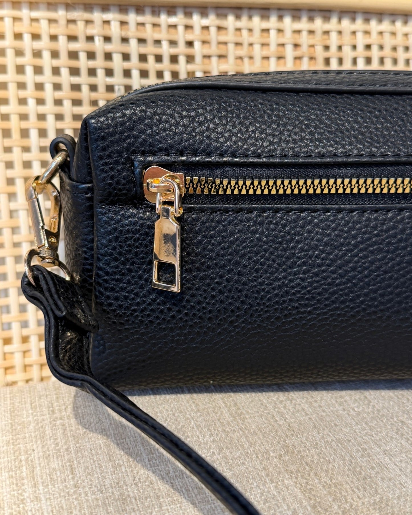 Close-up of a black pebbled leather handbag with a gold zipper and gold-tone hardware, resting on a neutral textured surface with a woven background.