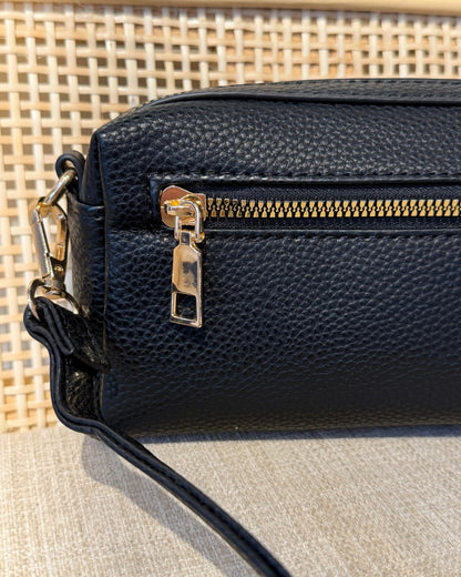 Close-up of a black pebbled leather handbag with a gold zipper and gold-tone hardware, resting on a neutral textured surface with a woven background.