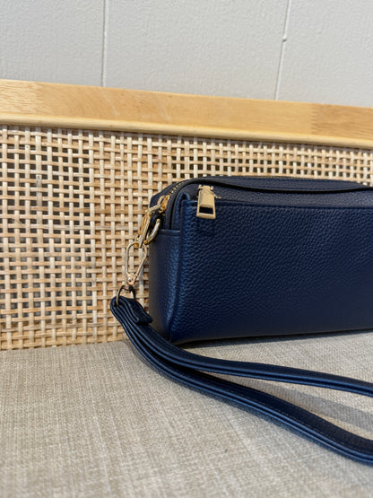 A navy blue pebbled leather wristlet bag with gold-tone hardware, resting on a textured beige fabric surface in front of a woven cane panel and light wood background.