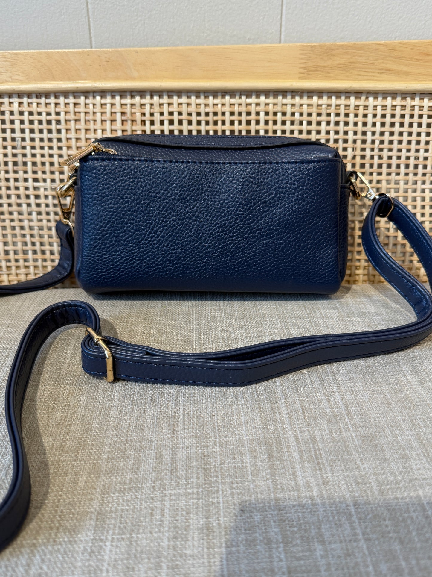 A navy blue pebbled leather crossbody bag with gold-tone hardware, displayed on a woven cane chair seat against a light gray wall.
