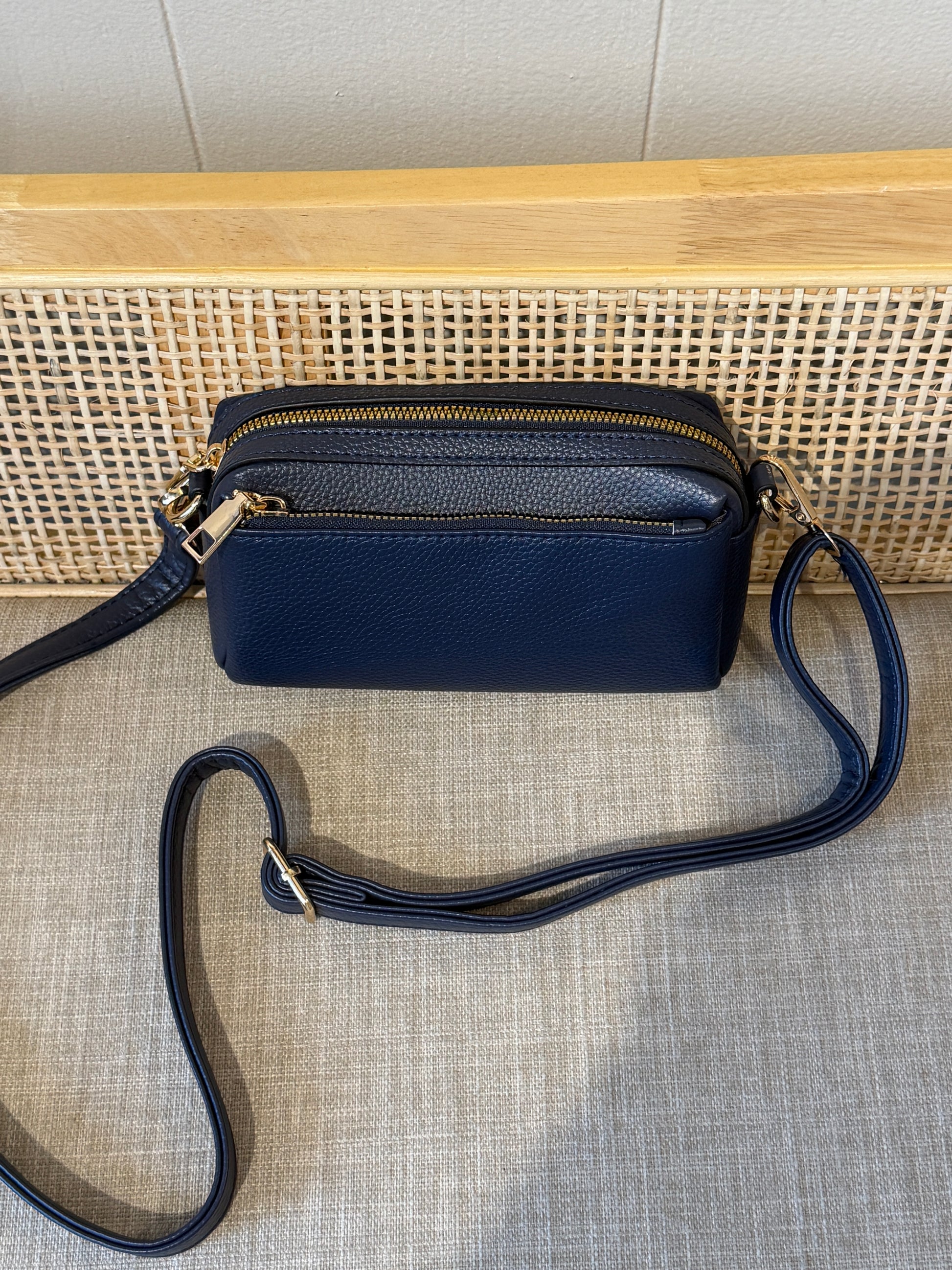 A navy blue pebbled leather crossbody bag with gold-tone hardware, resting on a textured gray fabric surface in front of a woven cane panel and light wood background.