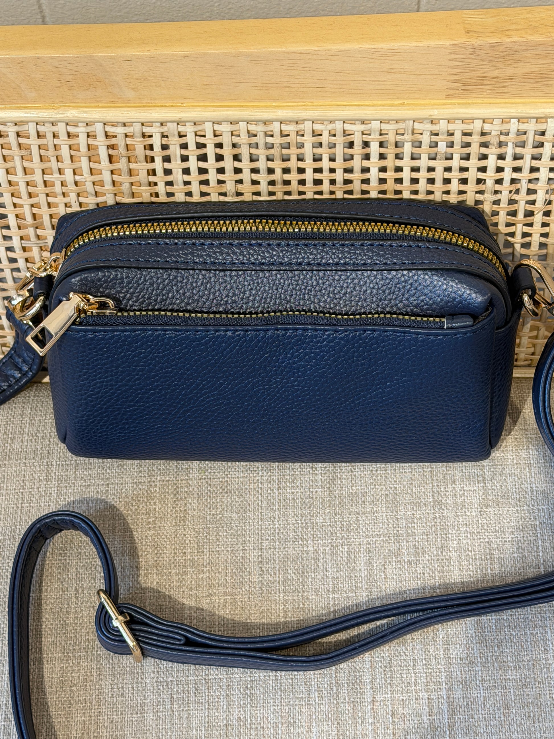 A navy blue pebbled leather crossbody bag with gold-tone zippers and hardware, displayed on a woven rattan surface with a matching strap in the foreground.