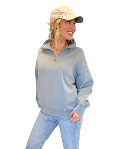 Woman smiling in jeans, cream baseball cap and Misty Blue Half-Zip Pullover with a relaxed fit, super-soft fabric, and hip-length hem, featuring a streamlined pocket-free design and adjustable half-zip neckline.