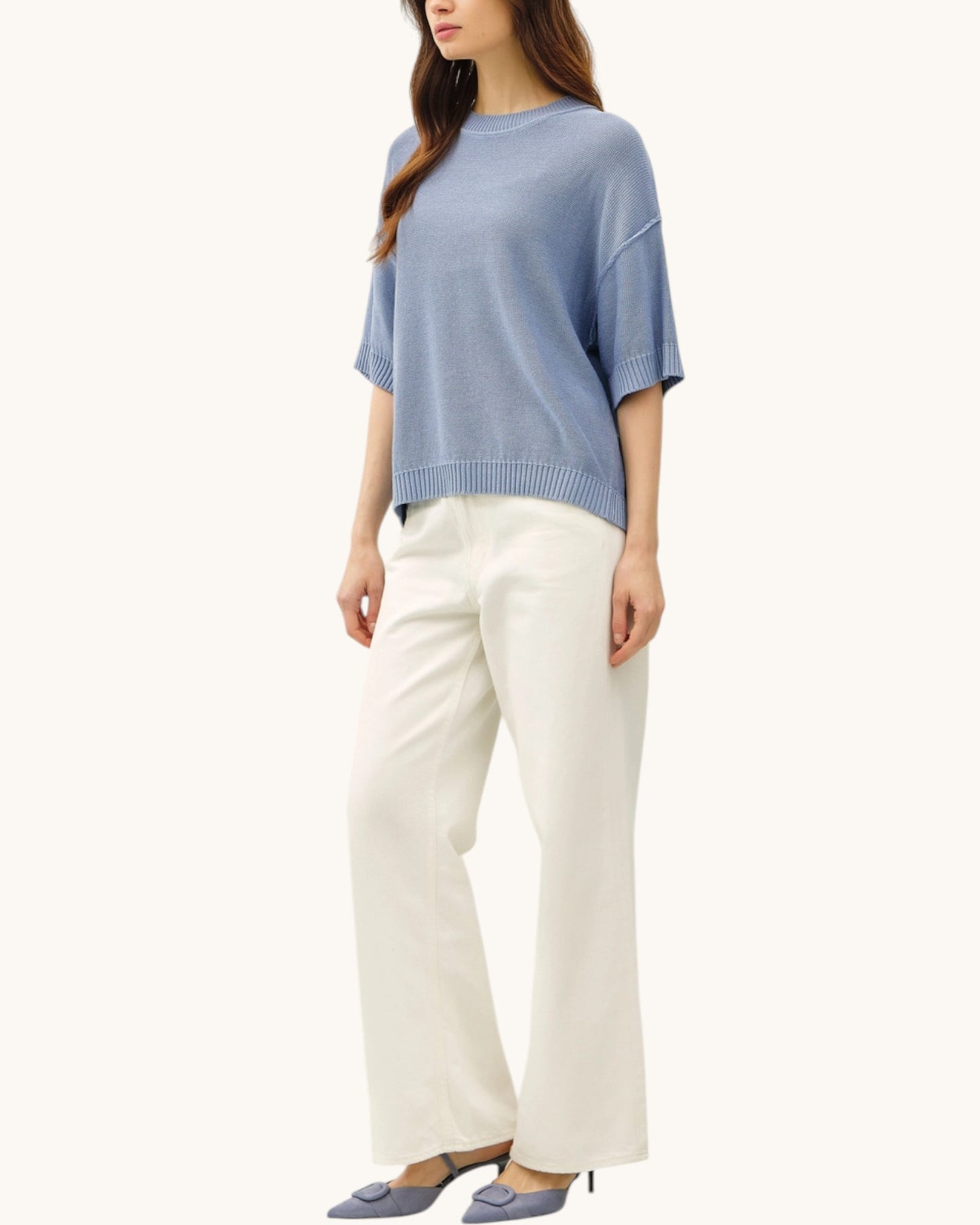 Woman wearing a blue lightweight sweater and white pants on a white background.